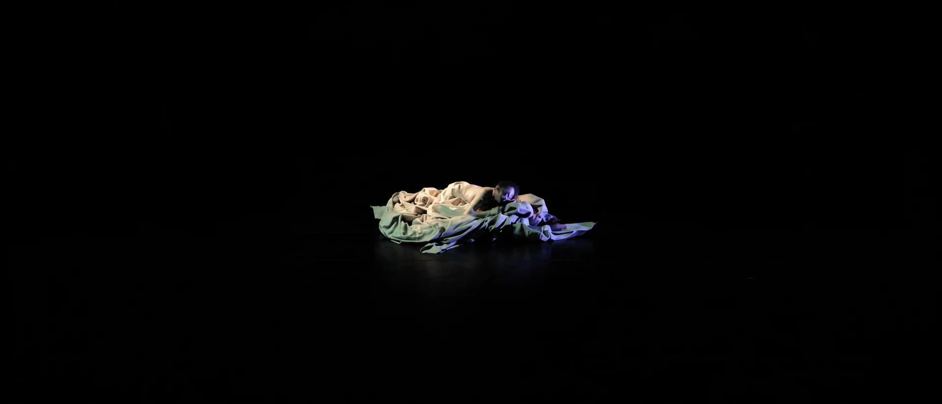 A wide dark stage with the dancer lying small at its centre, curled among the pale cloth.