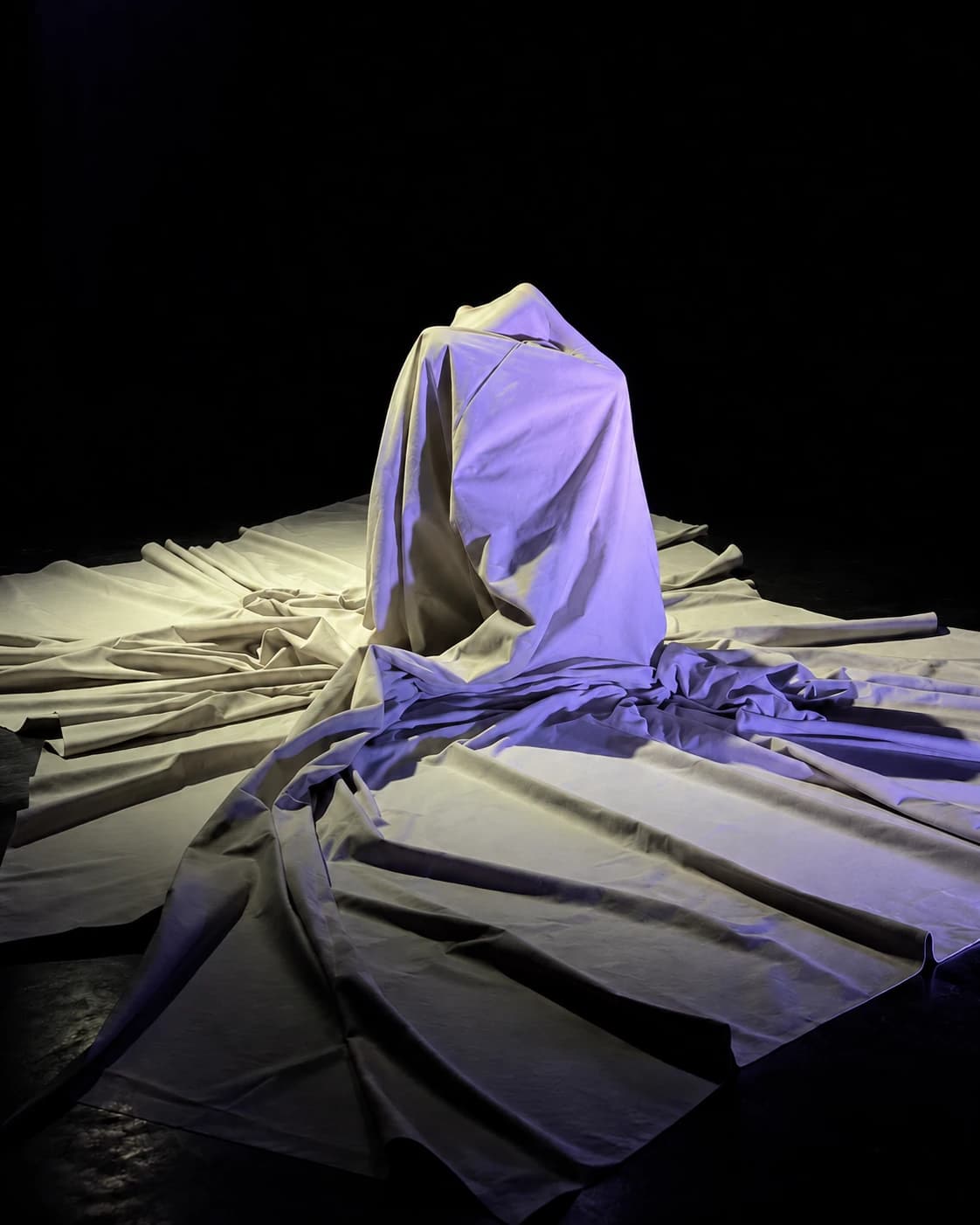 The dancer sits fully covered by the pale cloth, which spreads out from the body across the floor in broad folds under violet and pale green light.