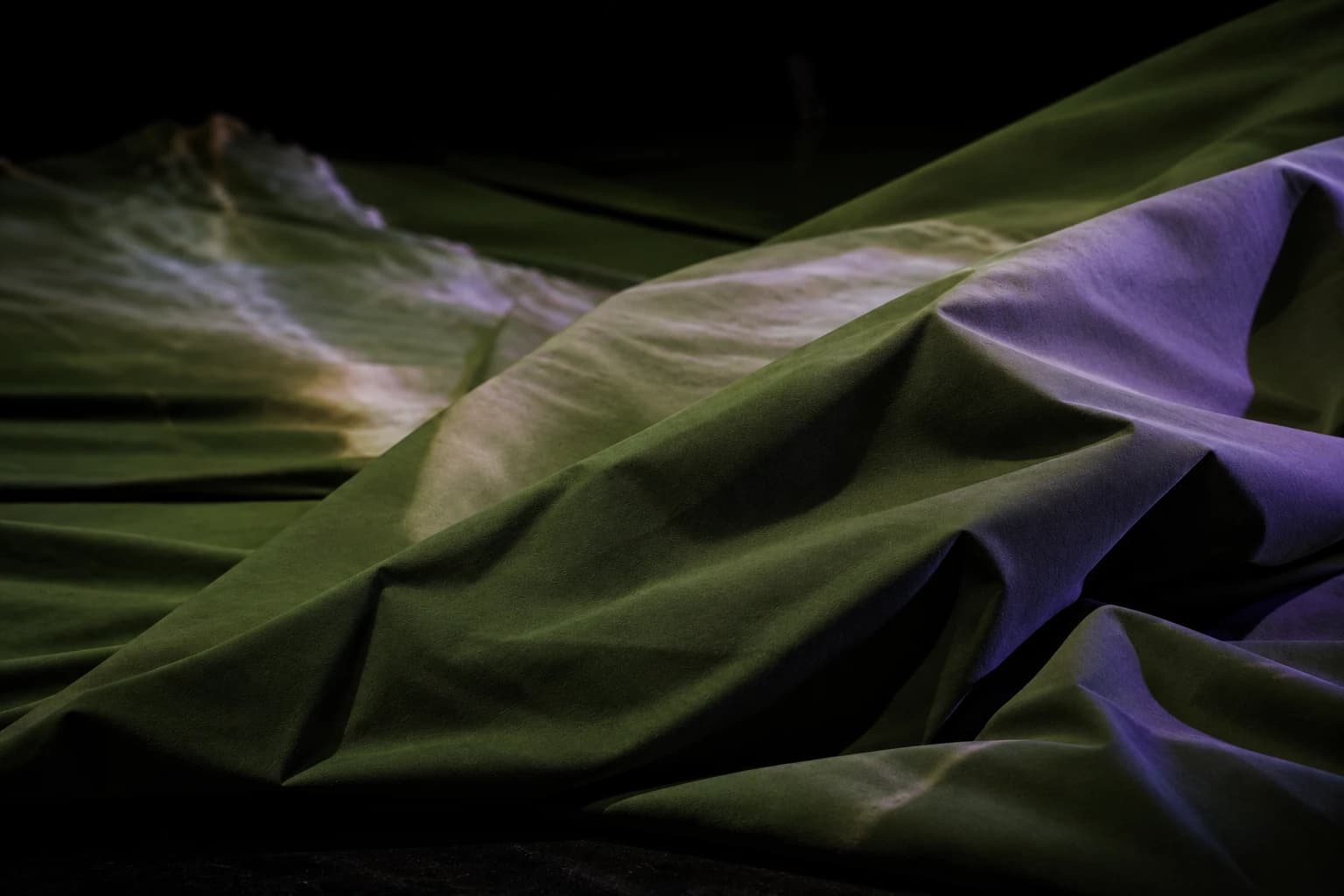 Close view of the pale cloth lying in long folds on the stage floor, washed in green and violet projected light.