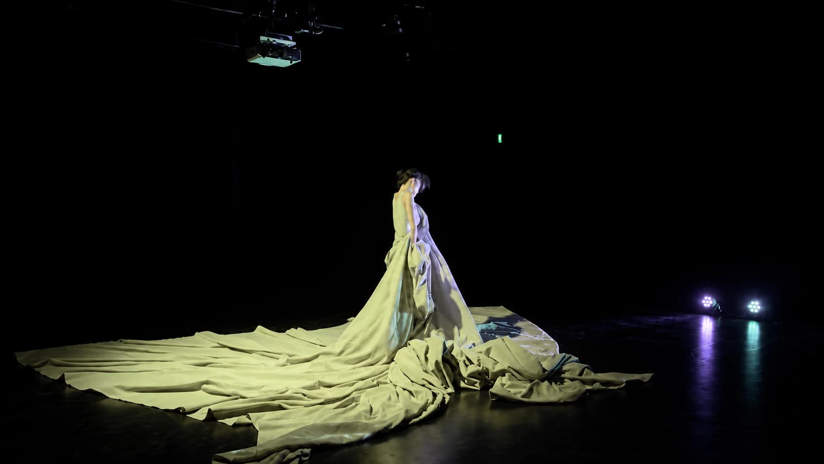 On a dark stage beneath a hanging projector, the dancer stands with head bowed, a large pale cloth gathered at the body and spreading far across the floor.