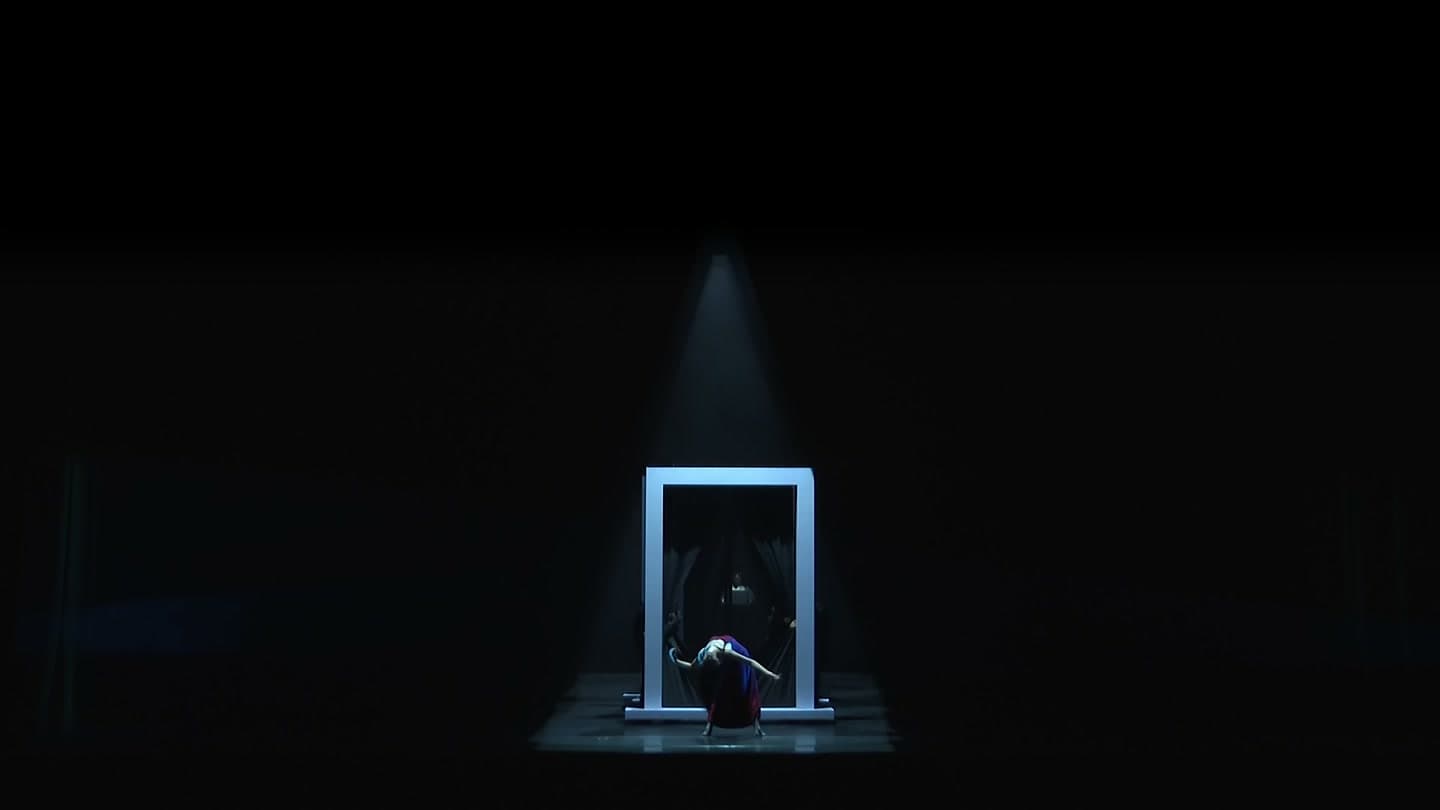 A single performer leans back in front of a white rectangular frame, lit from above in blue on an otherwise dark stage.
