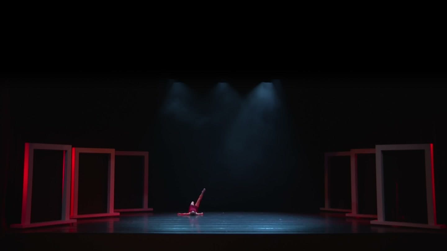 A performer lies on the floor of a dark stage, one leg raised, between rows of red-lit rectangular frames.