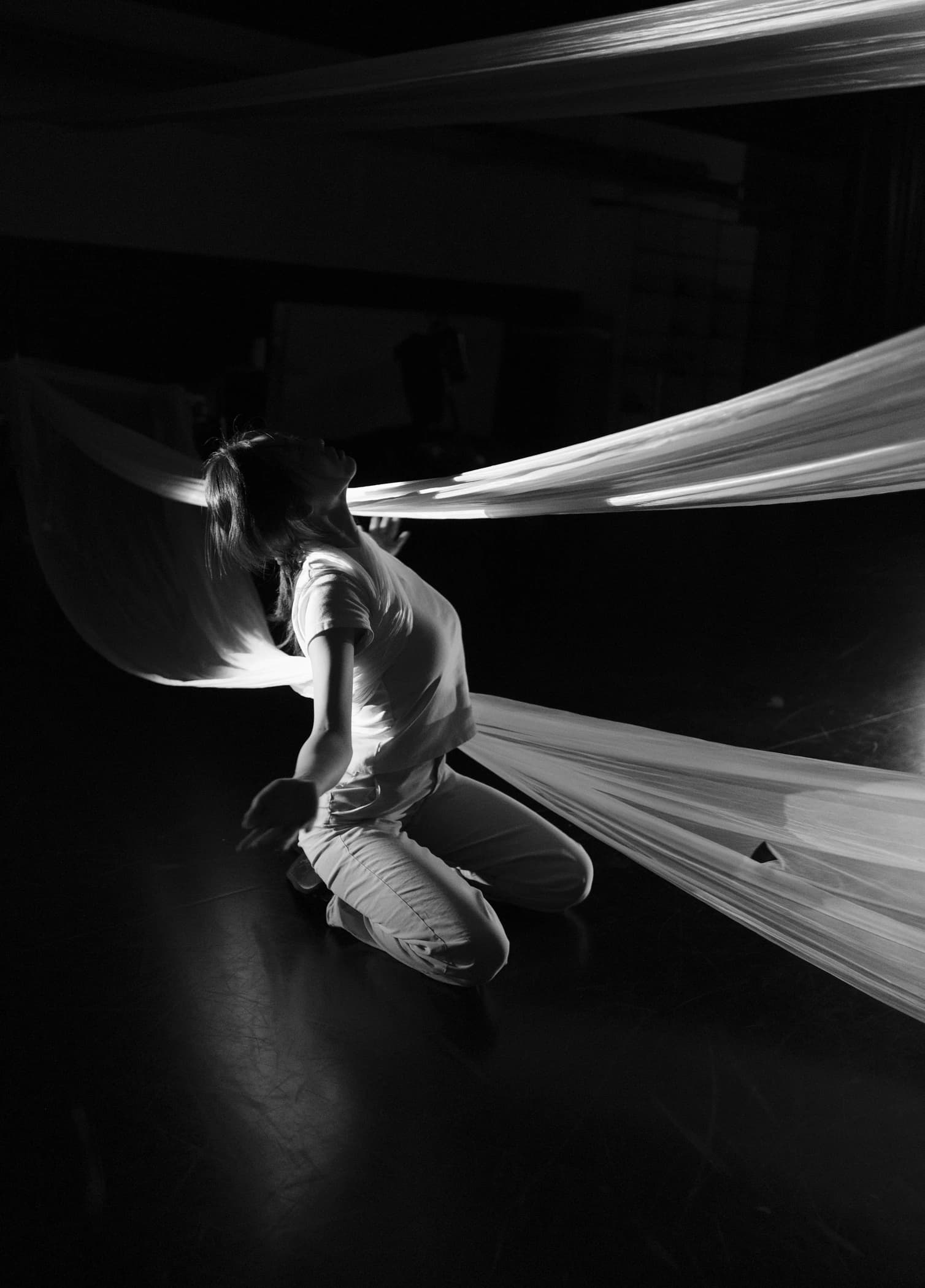 Black-and-white photograph: a performer in pale clothing kneels and arches back, face lifted toward broad translucent sheets that sweep past the body across a dark studio.